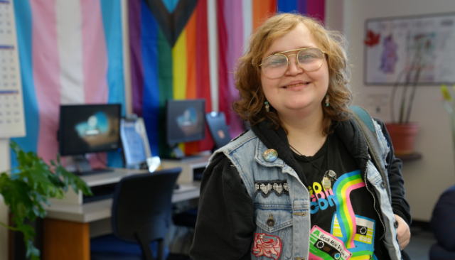 A student smiling and wearing cool clothes with various Pride flags in background