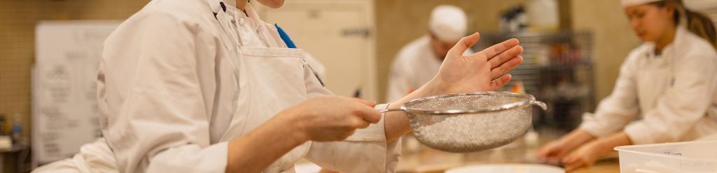 a culinary baking student with dyed blue hair and piercings carefully sprinkles powdered sugar on a desert