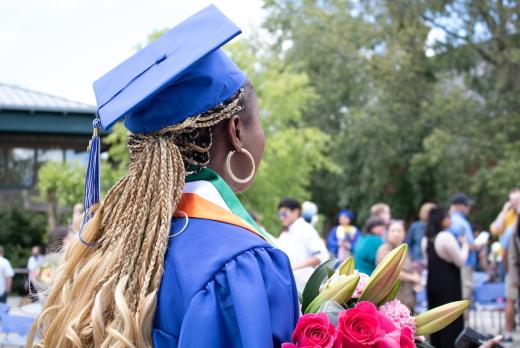 a black woman, an international student, in blue cap and gown turned from the camera, holding flowers and looking over the crowd at Commencement