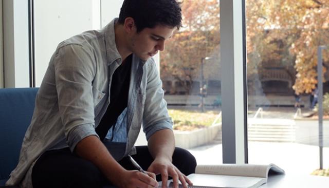 a young man sits by a window in the center building doing homework, with fall foliage outside