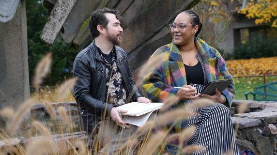 two students chat while holding notebooks and next to a fountain