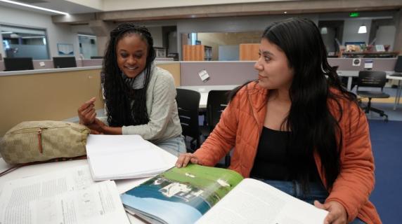 Two friends, young women in cozy winter jackets, studying in the library and smiling