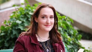 a young woman with long brown hair and maroon jacket reads a book on a bench