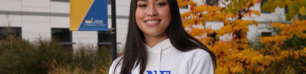 a young Asian american woman wearing a Lane Track and Field sweatshirt smiles at the camera with autumn foliage in the background