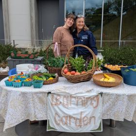 two women smiling at a Learning Garden table selling summer produce like cherry tomatoes and cucumbers